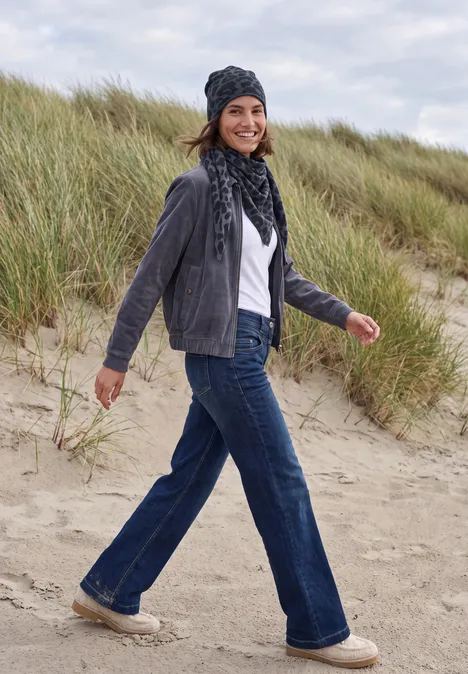 Une femme marche sur la plage devant des dunes, vêtue d’un jean, d’une veste grise, d’une écharpe et d’un bonnet.