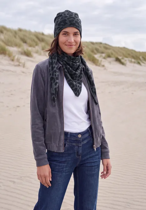 Une femme sur une plage de dunes porte un bonnet, une écharpe, une veste grise, un t-shirt blanc et un jean.