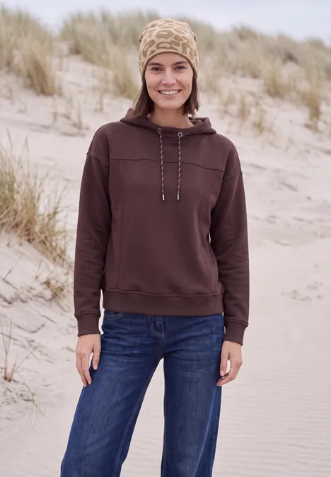 Femme en sweat à capuche marron et jean souriant sur une plage avec des dunes en arrière-plan.