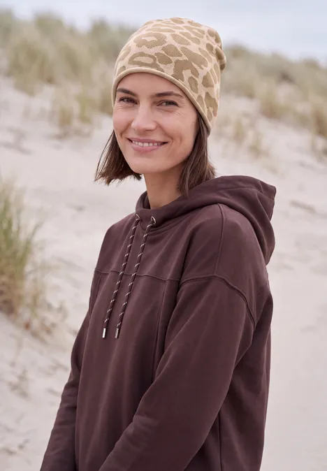 Une femme souriante en sweat à capuche marron et casquette beige à motifs girafe sur une plage.