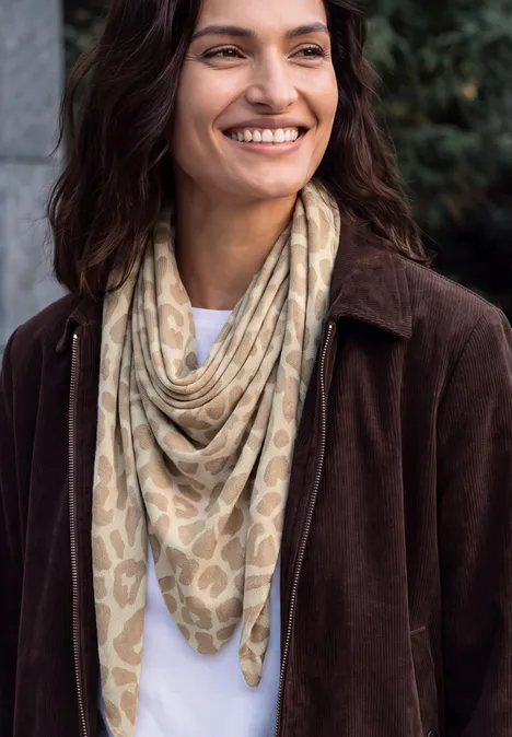 Femme en manteau marron et foulard beige à motifs sourit à l'extérieur.
