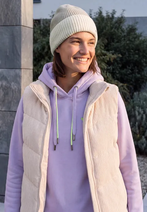 Femme souriante en bonnet beige, sweat violet clair et gilet devant un mur de pierre et des arbustes