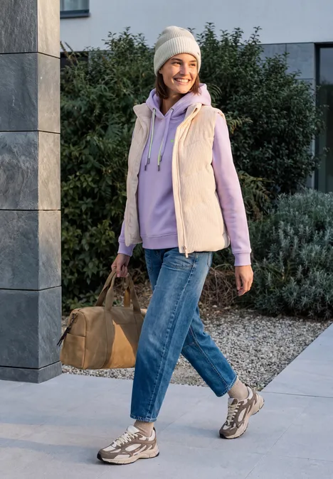 Femme joyeuse en bonnet, gilet et jean marche dehors avec un sac.