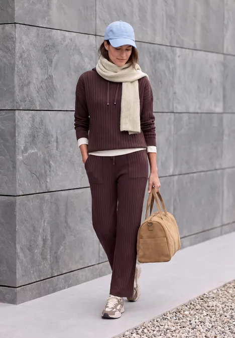 Femme en tenue décontractée marron, casquette et écharpe, marche avec un sac devant un mur gris.