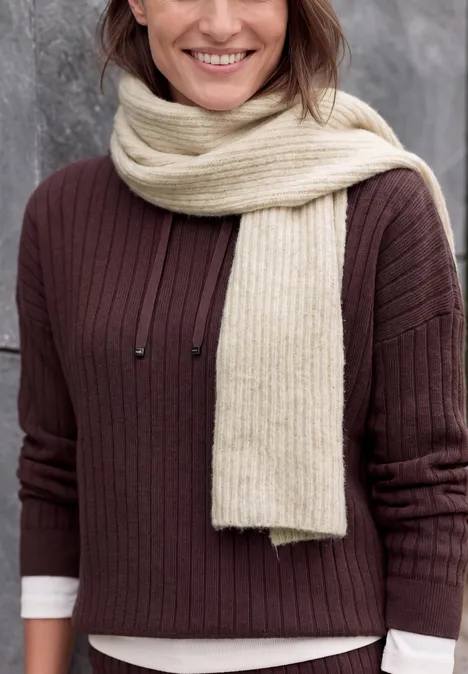 Une femme souriante en pull marron et écharpe crème devant un mur gris.