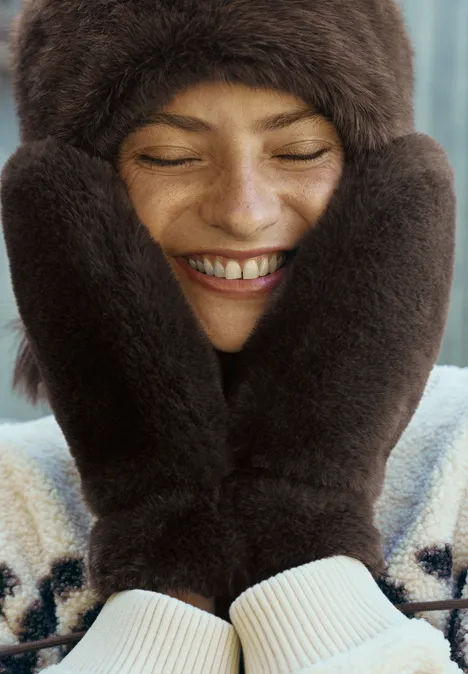 Warmtehandschoenen van imitatiebont in effen kleur chocolate sauce brown Eine lächelnde Frau mit Pelzmütze und Handschuhen hält sich die Hände an die Wangen.