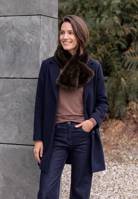 Fake Fur Schal in Unifarbe chocolate sauce brown Eine lächelnde Frau in Jeans, braunem Pullover, blauem Mantel und braunem Pelzschal steht draußen vor einer Wand.