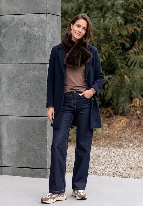 Fake Fur Schal in Unifarbe chocolate sauce brown Eine Frau in Mantel, Schal und Jeans steht vor einer Mauer und Grün, lächelt locker.
