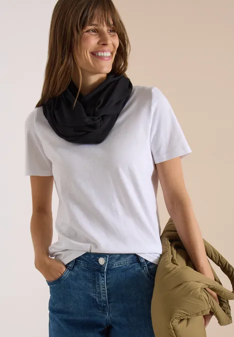 Effen Loop-sjaal Black Frau trägt weiße T-Shirt, Jeans und schwarzen Loop-Schal, hält eine olivgrüne Jacke und lächelt.