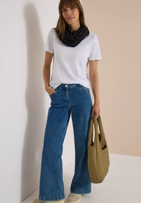 Effen Loop-sjaal Black Frau mit weißem T-Shirt, weitem Jeans, schwarzem Halstuch und grünem Beutel vor beiger Wand.