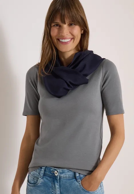 Effen Loop-sjaal urban dark blue Eine lächelnde Frau in grauem T-Shirt und dunkelblauem Schal, Hand in Hosentasche, vor hellem Hintergrund.