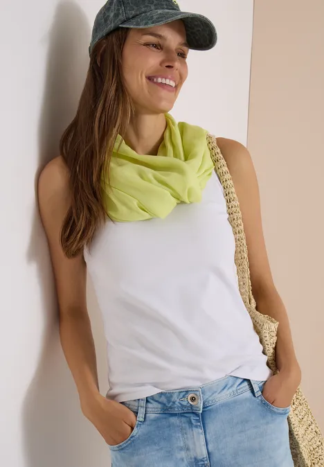 Effen Loop-sjaal citrin green Lächelnde Frau mit Cap, weißem Top, grünem Schal, Jeans und Strohtasche lehnt an einer Wand.