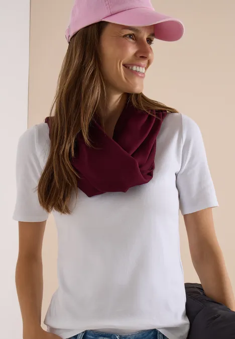 Effen Loop-sjaal red wine Eine lächelnde Frau in weißem T-Shirt, rosa Cap und weinrotem Schal über den Schultern, hält eine Jacke.