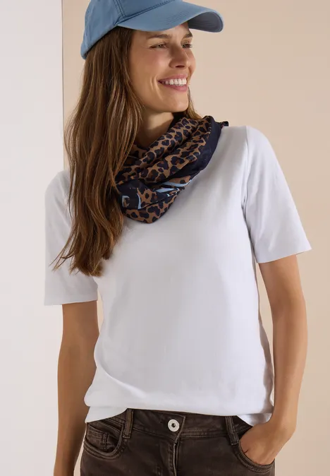 Loop-sjaal met luipaardprint creamy mocha Frau mit blauem Cap, weißem T-Shirt und gemustertem Schal, lächelnd mit einer Hand in der Tasche.