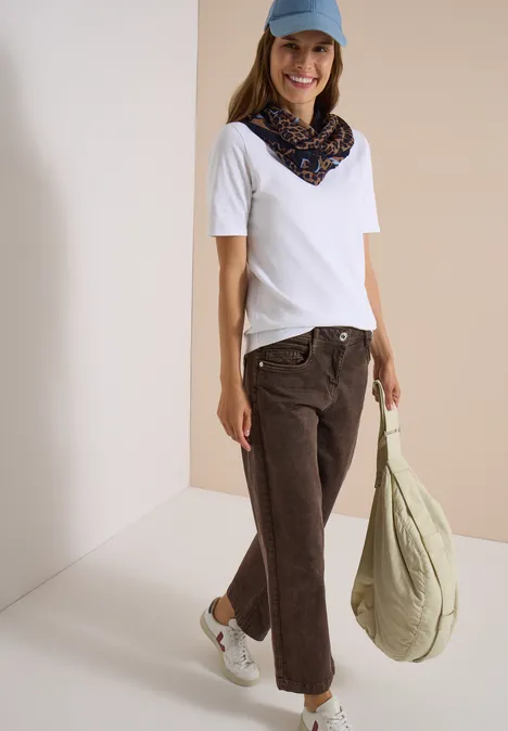 Loop-sjaal met luipaardprint creamy mocha Frau in weißem T-Shirt, braunen Jeans, blauem Cap und Halstuch trägt grüne Tasche und lächelt.
