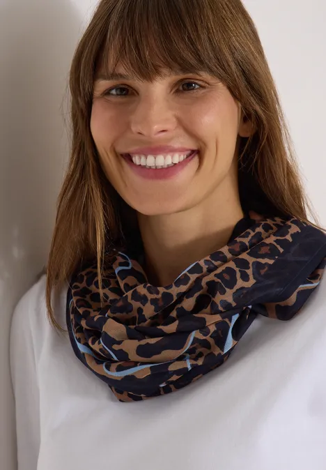 Loop-sjaal met luipaardprint creamy mocha Porträt einer lächelnden Frau mit braunen Haaren, weißem Oberteil und blau-braunem Tiermuster-Schal.