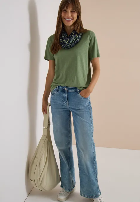Loop-sjaal met luipaardprint dried olive green Frau in grünem T-Shirt, weitem Jeans, Halstuch und großer beiger Tasche vor neutralem Hintergrund.