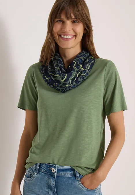 Loop-sjaal met luipaardprint dried olive green Eine lächelnde Frau trägt grünes T-Shirt und gemusterten Schlauchschal vor hellem Hintergrund.