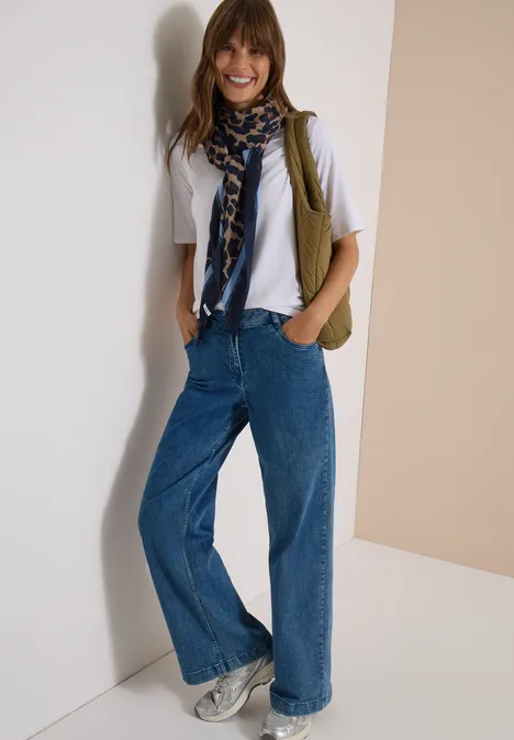 Frau in weitem Jeans, weißem T-Shirt, Schal und Sneakers posiert lächelnd vor neutralem Hintergrund.