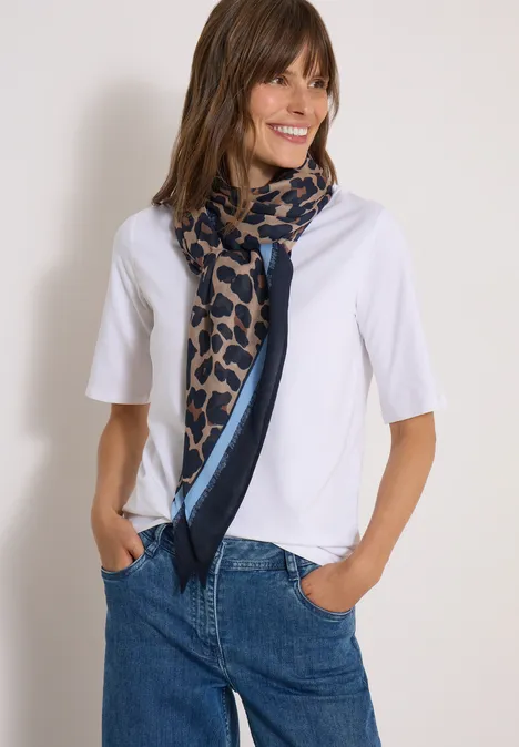 Kundin trägt weißes Shirt, gemusterten Schal und Jeans, lächelnd mit Händen in den Taschen.