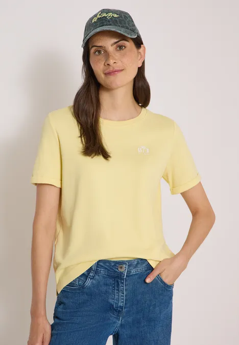 Eine Frau mit gelbem T-Shirt, blauer Jeans und Baseballcap lächelt vor neutralem Hintergrund.