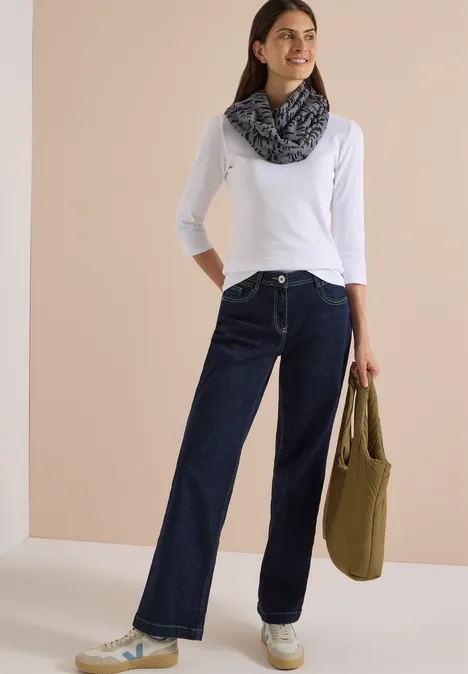 Frau in weißem Langarmshirt, dunklen, weit geschnittenen Jeans, gemustertem Schal und olivgrüner Tasche vor neutralem Hintergrund.
