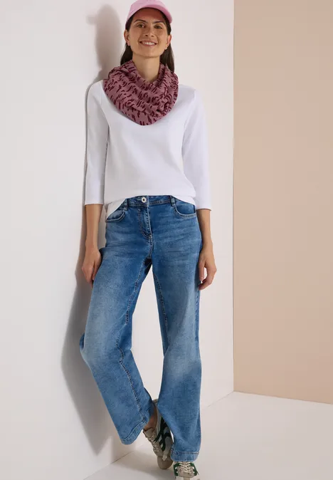 Frau in Jeans, weißem Shirt, rosa Mütze und Schal lehnt lächelnd an heller Wand.