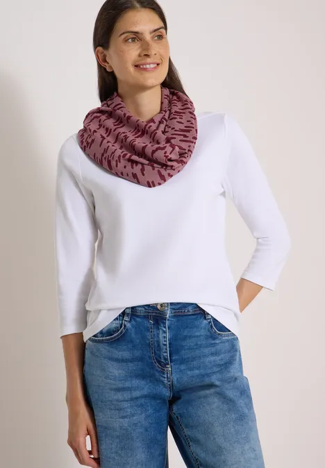Frau in weißem langärmligem Shirt, blauen Jeans und rosafarbenem Loop-Schal mit schwarzem Muster.