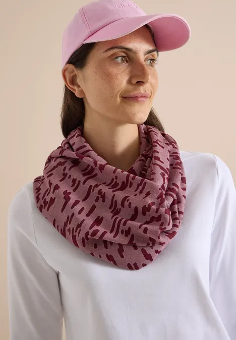 Frau mit rosa Cap und gemustertem Schlauchschal, weißes Shirt, neutraler Hintergrund.