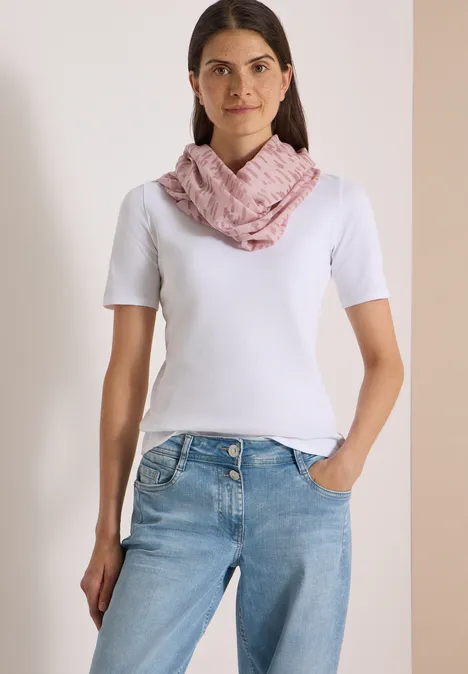 Eine Frau trägt ein weißes T-Shirt, Jeans und einen rosa Schal, steht vor hellem Hintergrund.