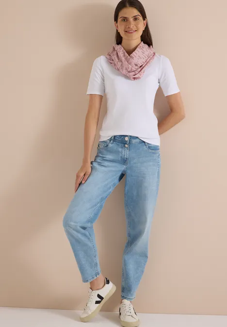 Frau in weißem T-Shirt, blauen Jeans, rosa Schal und weißen Sneakern vor neutralem Hintergrund.