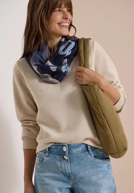 Een glimlachende vrouw met beige trui, jeans, blauwe sjaal en groene gewatteerde tas.