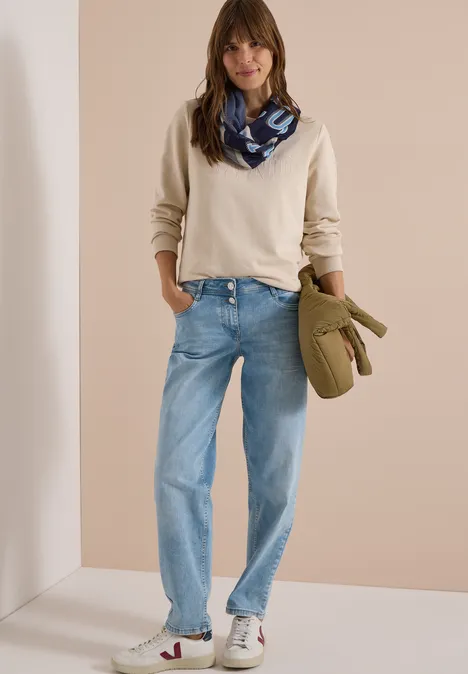 Vrouw in crèmetint trui, blauwe sjaal, losse jeans en witte sneakers, met groene tas.
