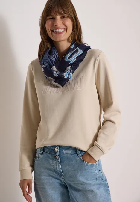 Vrouw in beige trui, spijkerbroek en blauwe sjaal lacht.