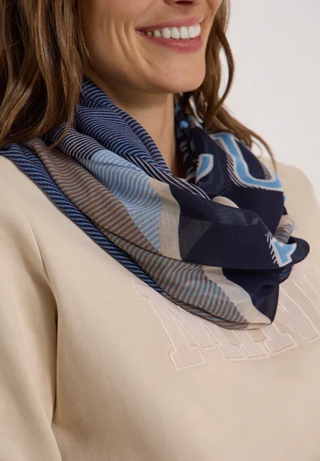 Vrouw met blauw dessin sjaal om de nek, beige trui en subtiele glimlach.
