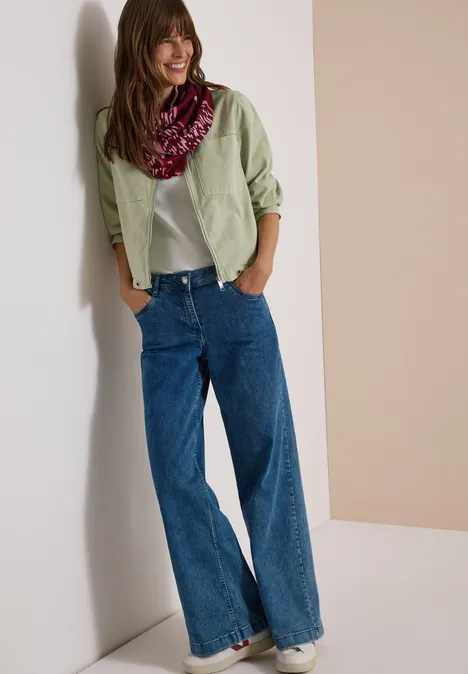 Vrouw in lichtgroen overhemd, wit shirt, wijde blauwe jeans en kleurrijke sjaal, ontspannen tegen de muur leunend.