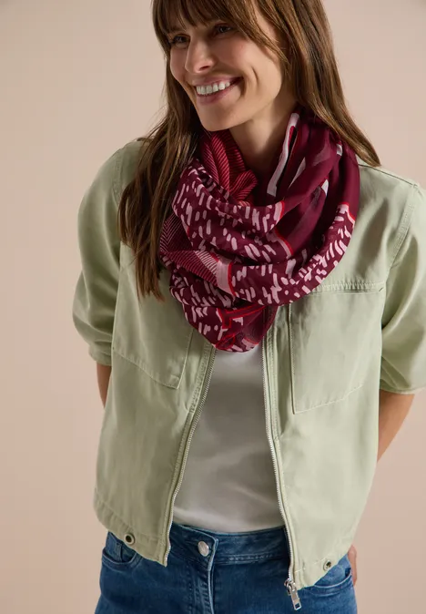 Vrouw met lichtgroen jasje, witte top, spijkerbroek en bordeauxroze sjaal met dessin, glimlachend.
