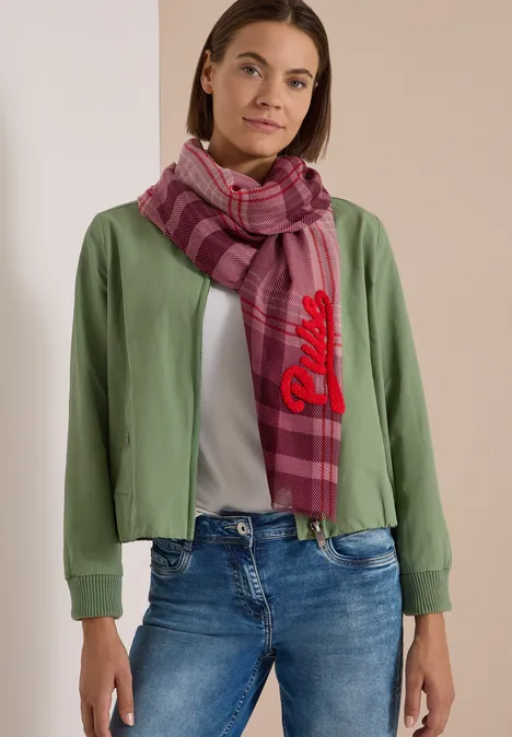 Frau trägt grünen Blouson, weißes Shirt, Jeans und karierten rosa-pinken Schal.