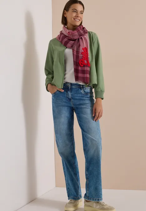 Frau in grüner Strickjacke, lila kariertem Schal und lockeren blauen Jeans vor neutralem Hintergrund.