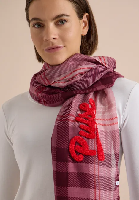Frau trägt weißen Pullover und rosa-lila karierten Schal mit roter 'Dune'-Stickerei.