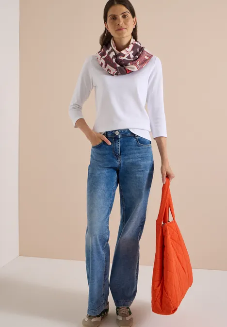 Frau in weißem 3/4-Armshirt, weiten Jeans und gemustertem Schal, mit orangefarbener Tasche.