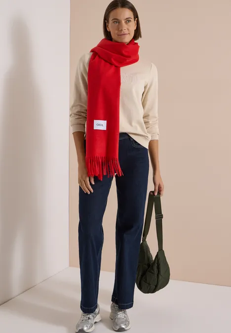 Frau trägt beigen Pullover, blaue Jeans, roten Schal und hält grüne Tasche.