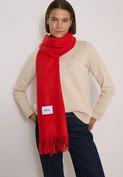 Frau mit rotem Schal, beigeem Pullover und Jeans vor neutralem Hintergrund.