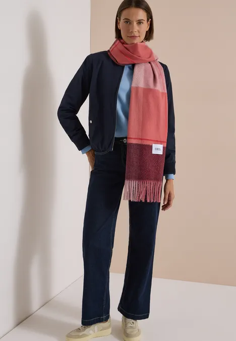 Vrouw draagt kleurblok-sjaal, marineblauw jasje, lichtblauwe top en wijde jeans voor neutrale achtergrond.