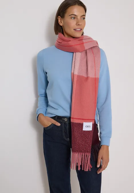 Vrouw met lichtblauwe trui, spijkerbroek en rood-roze sjaal, glimlachend met hand in zak.