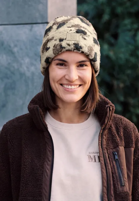 Bonnet en fausse fourrure à motif vache pearl beige mel. Jeune femme souriante en bonnet camouflage et veste polaire brune.