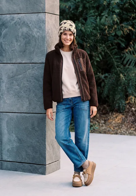 Bonnet en fausse fourrure à motif vache pearl beige mel. Femme souriante en bonnet, veste et jean appuyée contre une façade moderne en plein air.
