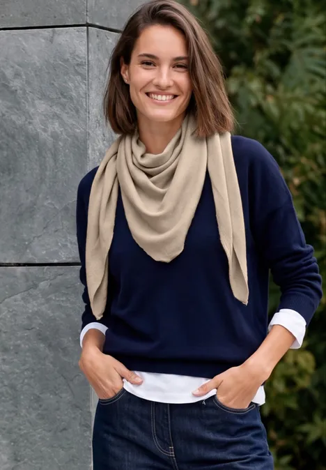 Eine lächelnde Frau im Freien trägt Jeans, weißes Hemd, dunkelblauen Pullover und beigen Schal.