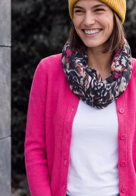 Écharpe Loop à imprimé graphique truffle brown Une femme souriante porte un bonnet jaune, une écharpe colorée et un cardigan rose.