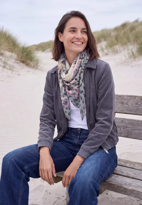 Tuch im Ausbrenner-Look truffle brown Eine Frau sitzt entspannt auf einer Strandbank, lächelt, trägt Jeans, Schal und Jacke vor Dünen.